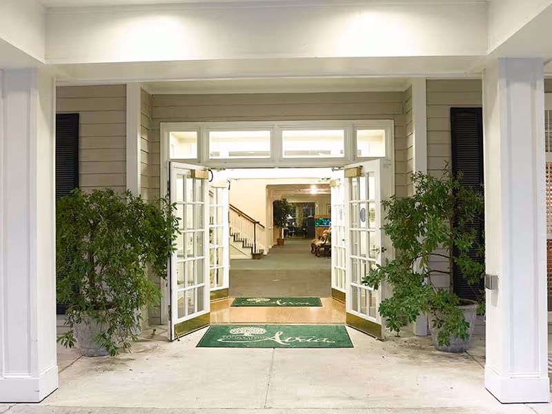 Entrance to a building with double glass doors open, flanked by two large potted plants. Inside, a hallway with carpet leads to a staircase and seating area. Green mats with white text are placed at the entrance.
