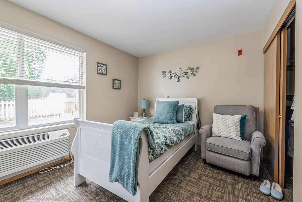 A cozy bedroom with a white single bed adorned with teal and patterned bedding, a matching teal throw blanket, and pillows. Next to the bed is a white nightstand with a teal lamp. A gray armchair with a white knitted pillow sits beside a wooden closet with sliding doors. A pair of slippers is placed on the carpeted floor near the armchair. A large window with white blinds lets in natural light, and the walls are decorated with small framed pictures and a metal floral wall art.