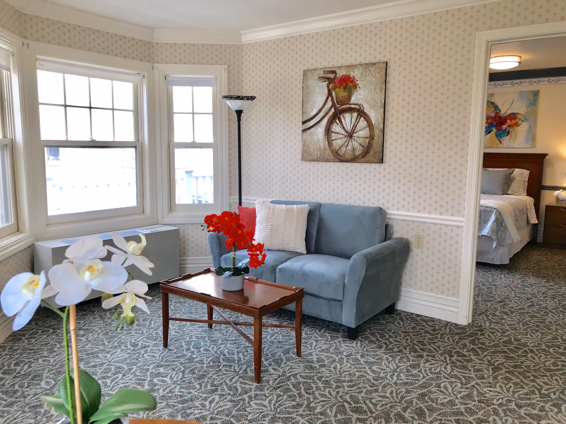 A cozy living room area with a light blue loveseat, a wooden coffee table with a red flower arrangement, and a floor lamp. The room has patterned carpet and wallpaper with a bicycle painting on the wall. Large windows allow natural light to fill the space. A bedroom with a bed and colorful artwork is visible through an open doorway.