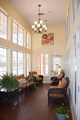 A bright indoor seating area with large windows on one side letting in natural light. The space features wicker chairs with striped cushions and decorative pillows, a hanging chandelier light fixture, potted plants, and a floral painting on the wall above a glass door leading outside.