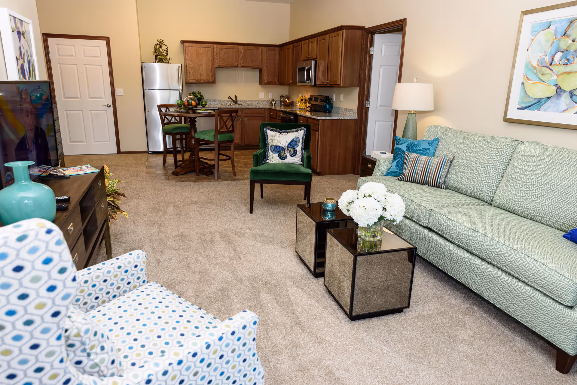 A cozy living room and kitchen area in a retirement community apartment. The living room features a light green sofa with decorative pillows, a patterned armchair, two mirrored coffee tables with a vase of white flowers, and a floor lamp. The kitchen has wooden cabinets, a stainless steel refrigerator, microwave, and stove. A small round dining table with four chairs is positioned near the kitchen. The walls are painted beige, and there is a colorful framed artwork above the sofa.