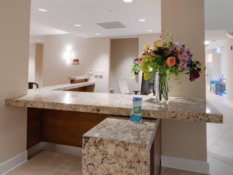 Reception desk area with a granite countertop, a vase of colorful flowers, and a hand sanitizer dispenser. The background shows a hallway with seating and soft lighting on beige walls.