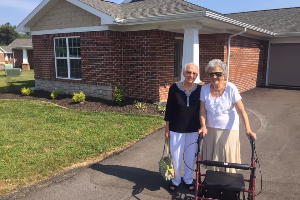 Two older women, one using a walker, stand on a driveway in front of a single-story brick house.