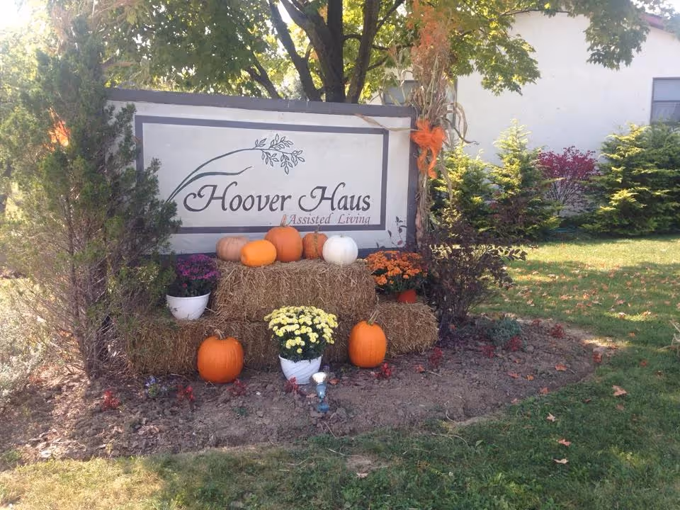 Outdoor sign reading "Hoover Haus Assisted Living" decorated with pumpkins, hay bales and potted flowers under a tree.