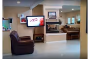 Interior view of a common area in an assisted living facility featuring a brown recliner chair, a wall-mounted flat screen TV displaying a red and white screen, a double-sided fireplace, a piano in the background, and a fish tank on the left side.
