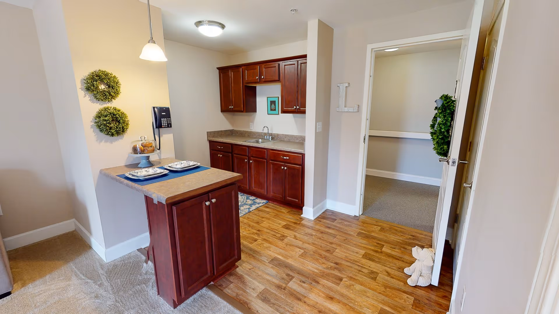 Interior view of a small kitchen area with wooden cabinets and a countertop island set with two plates and napkins. The floor is a combination of wood and carpet. There are two green wreaths hanging on the wall, a wall-mounted phone, and an open door leading to another room with a teddy bear placed on the floor near the door.