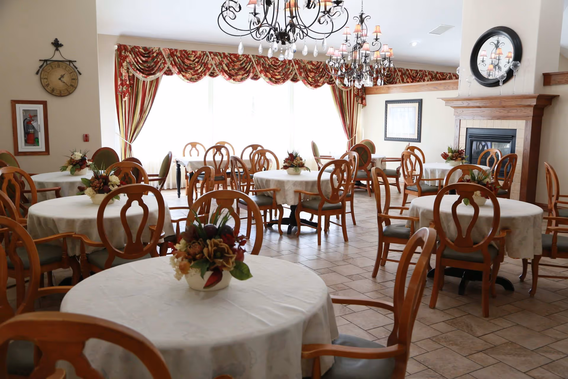 Bright dining room with round tables covered in white tablecloths, wooden chairs, floral centerpieces, chandeliers, and a fireplace.
