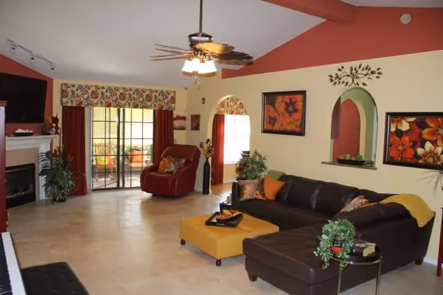 A spacious living room with a high vaulted ceiling painted red on one side. The room features a dark brown sectional sofa with colorful throw pillows and a yellow ottoman in front. There is a red armchair near sliding glass doors that lead to an outdoor patio. The walls are decorated with floral artwork and a decorative metal wall piece above an arched pass-through window. A ceiling fan with lights hangs from the ceiling, and there are plants placed around the room, including near a fireplace on the left side.