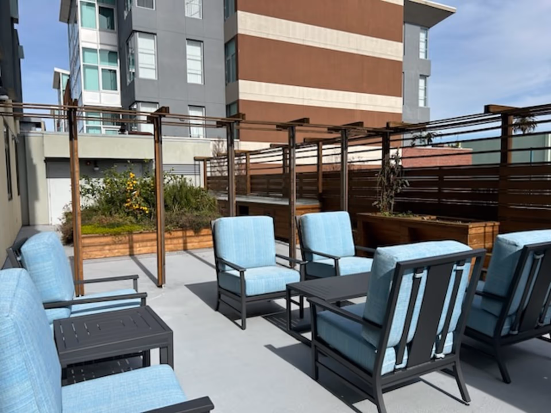 Outdoor patio area with light blue cushioned chairs arranged around a small black table. There are wooden planters with greenery and a wooden pergola structure. The background shows a multi-story building with windows.
