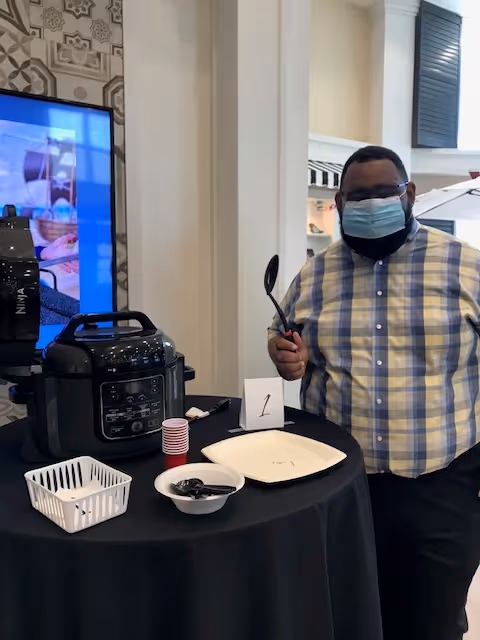 A man wearing a face mask and a yellow and blue plaid shirt stands next to a round table covered with a black tablecloth. On the table are a black Ninja air fryer, a white basket, a bowl with black spoons, a stack of small red cups, and a white plate with a number 1 card. The background shows part of an interior space with white walls and a large screen.