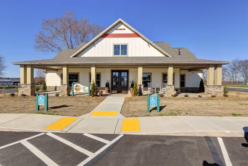 Front exterior view of Cedar Springs Independent Living facility, showing a single-story building with a covered porch supported by columns, wheelchair accessible ramps, and a clear blue sky in the background.