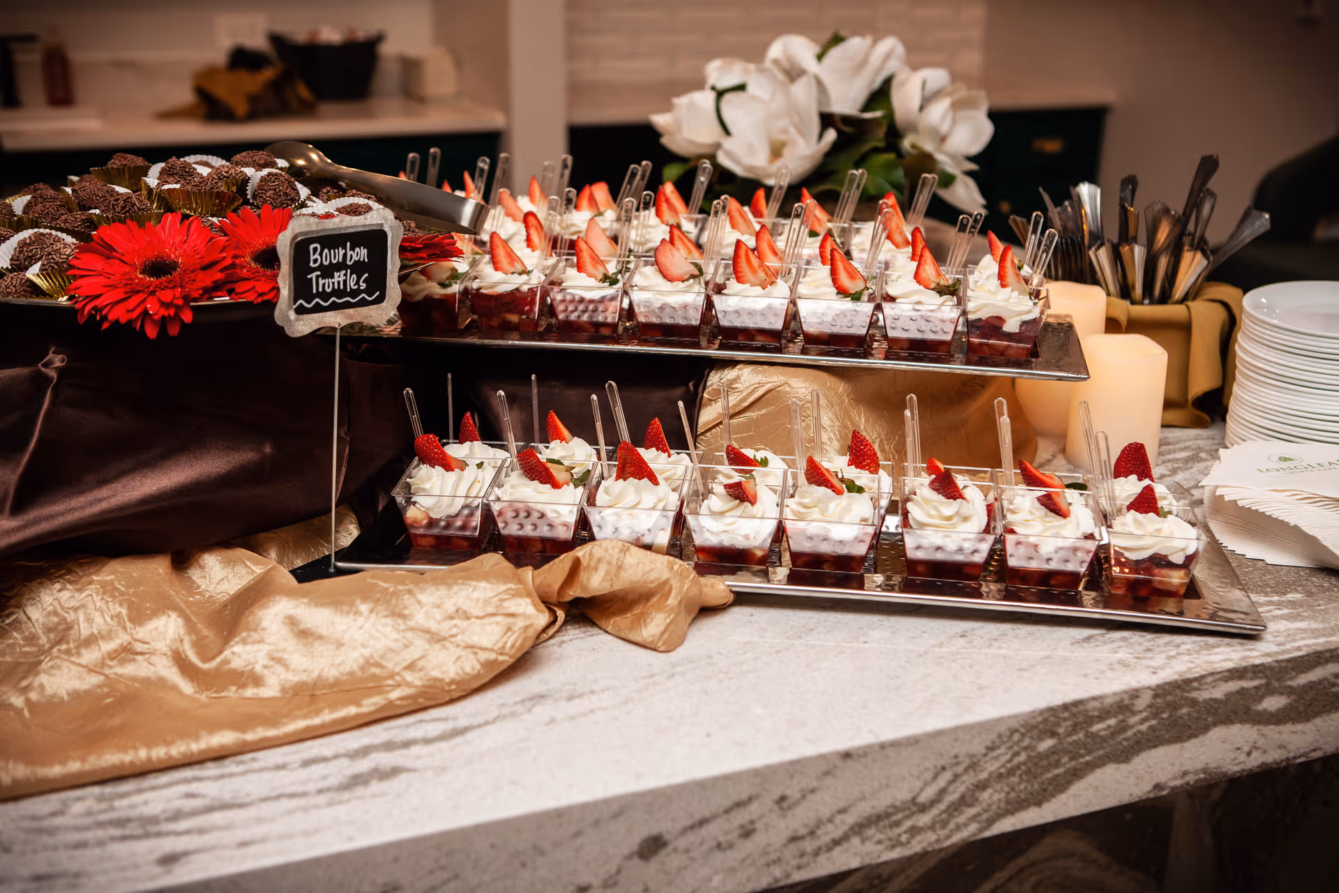 Tray of individual strawberry‑topped dessert cups arranged on a buffet table with plates and utensils nearby.