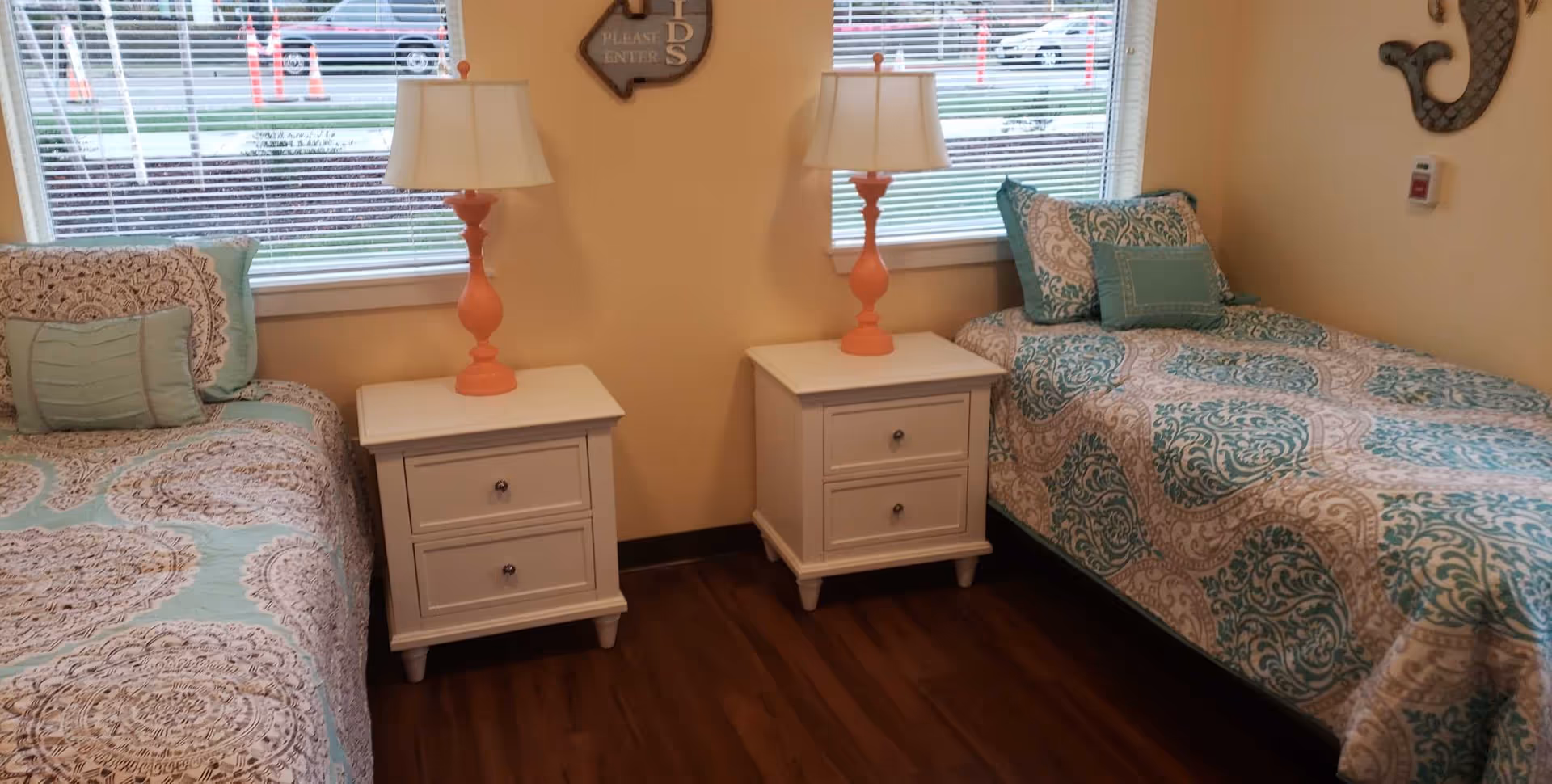 A bedroom with two twin beds separated by two white nightstands, each with an orange lamp. The beds have patterned bedspreads in teal, beige, and white colors with matching pillows. Two windows with blinds are behind the nightstands, and a wall decoration is visible on the right side.