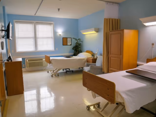 A bright hospital or rehabilitation room with two adjustable beds, each with white linens and wooden headboards and footboards. The room has light blue walls, a window with blinds, a wooden cabinet, a striped armchair, a potted plant, and a wall-mounted TV. There is a privacy curtain partially drawn between the beds.