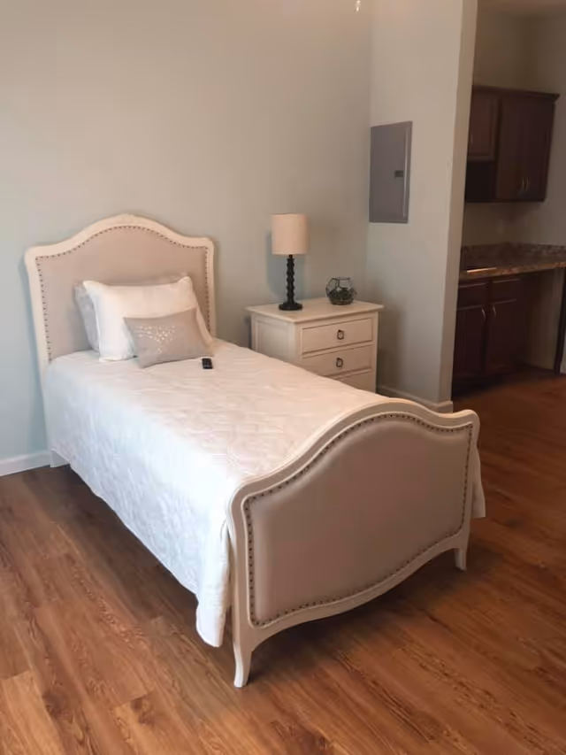 A neatly made single bed with a white quilt and pillows in a room with wooden flooring. Next to the bed is a white nightstand with two drawers, a black lamp with a beige shade, and a small decorative item. In the background, there is a partial view of a kitchen area with dark wooden cabinets and a granite countertop.