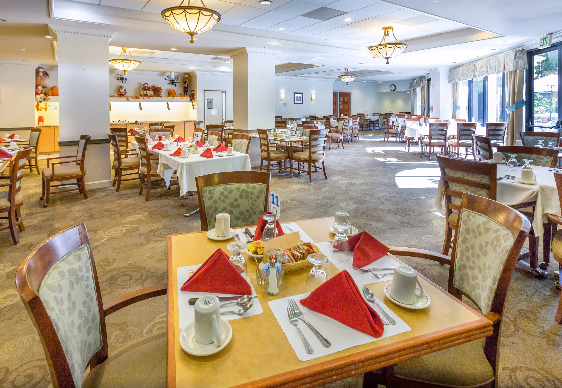 A spacious dining room in a senior living facility with multiple tables set for meals. Each table has white tablecloths, red folded napkins, cups, glasses, and silverware. The room is well-lit with ceiling lights and natural light coming through large windows on the right side. The decor includes light-colored walls, patterned carpet, and autumn-themed decorations on a shelf in the background.