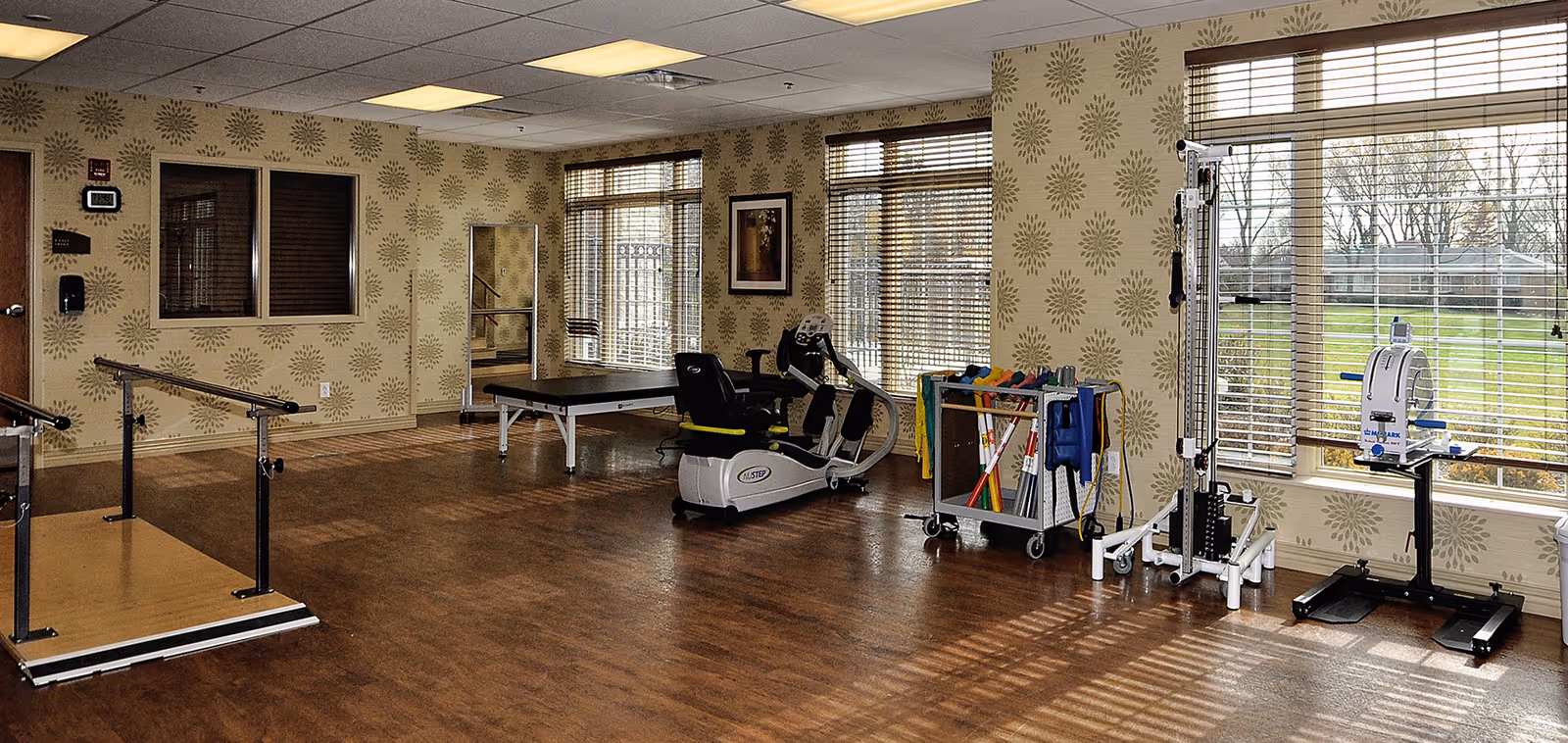 A spacious rehabilitation or physical therapy room with wooden flooring and patterned wallpaper. The room contains parallel bars for walking exercises, a recumbent exercise bike, a treatment table, and various exercise equipment near large windows with blinds. Outside the windows, a grassy area and trees are visible.