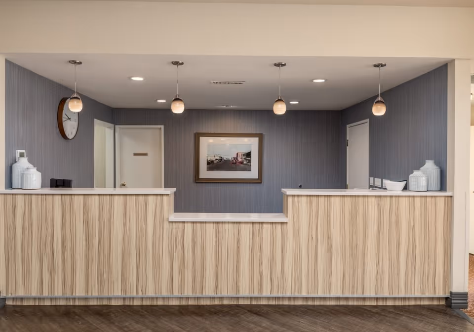 Reception desk area in a senior living facility with a light wood panel front, three pendant lights hanging from the ceiling, a wall clock on the left wall, a framed picture on the back wall, and two doors behind the desk.