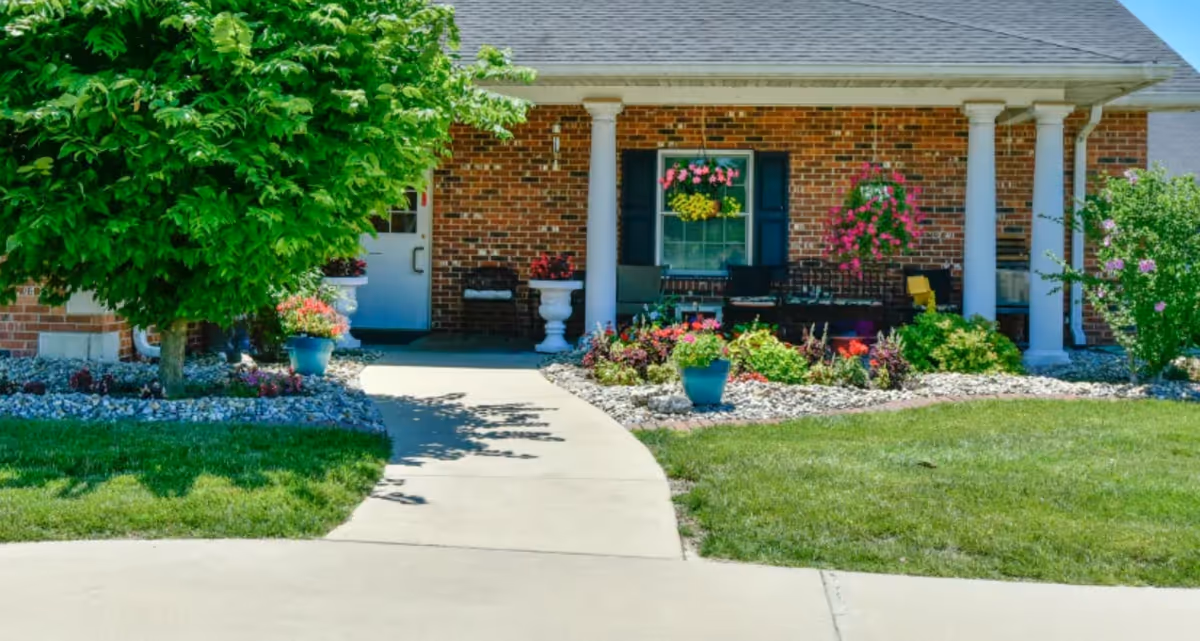 Front entrance of a brick building with a curved concrete walkway, porch columns, hanging baskets and potted flowers, and a manicured lawn.