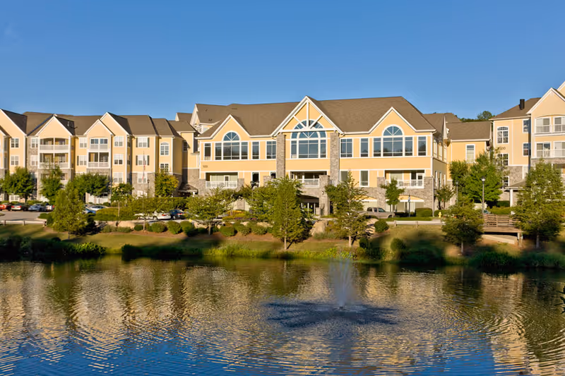 Exterior view of a large senior living facility named Park Springs with multiple stories, large windows, and a pond with a water fountain in the foreground. The building is surrounded by trees and landscaping under a clear blue sky.