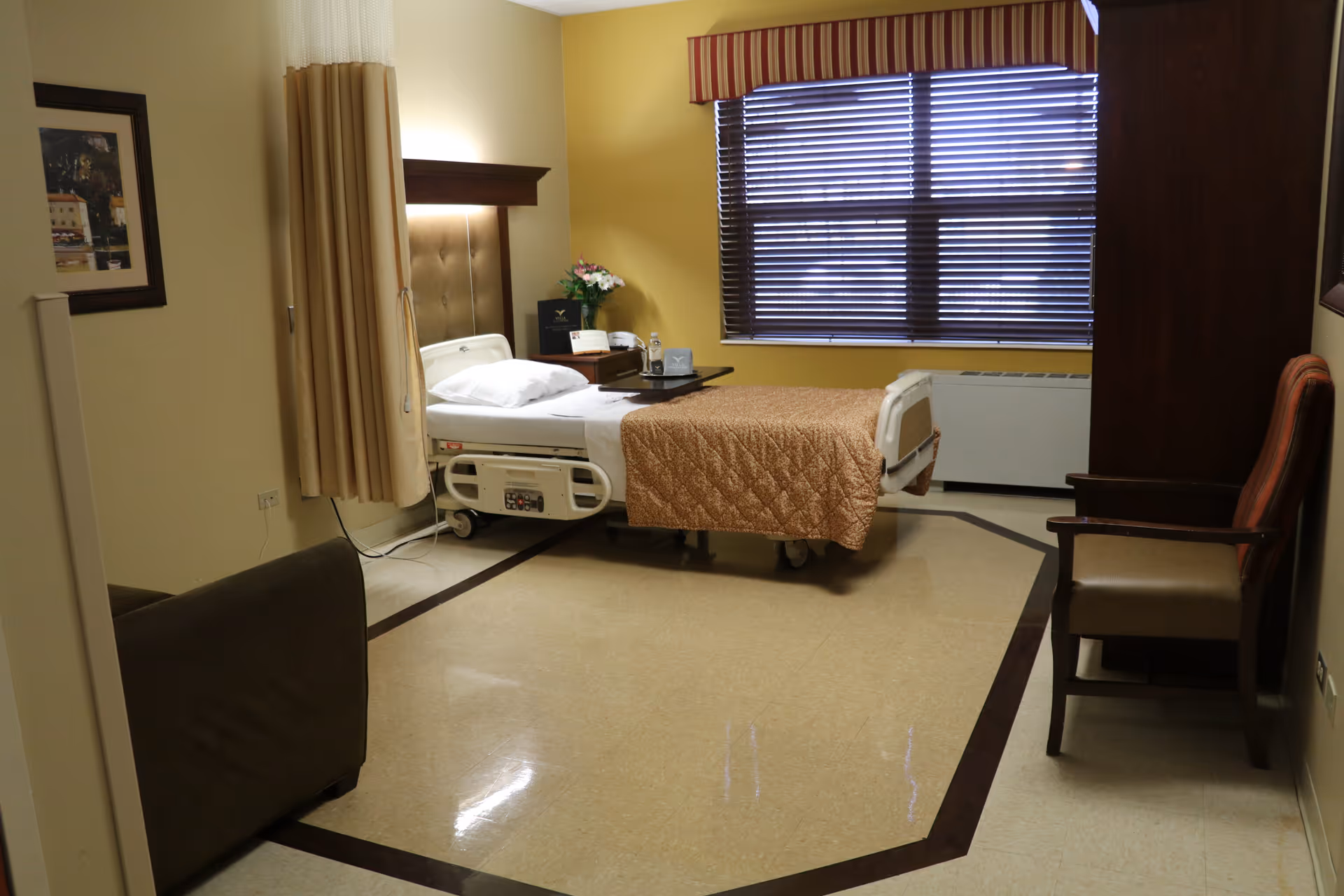 A furnished senior care bedroom featuring an adjustable hospital bed with a bedside table, chairs, and a window with blinds.