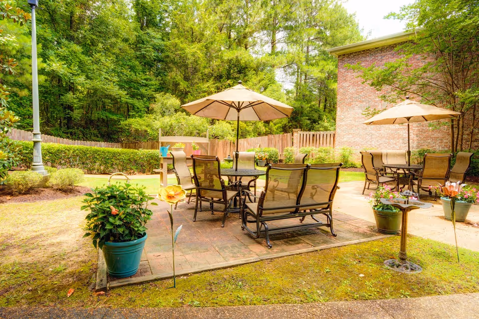 Outdoor patio area with two sets of metal tables and chairs, each shaded by large beige umbrellas. The patio is surrounded by green plants, potted flowers, and a wooden fence with trees in the background. A brick building wall is visible on the right side.