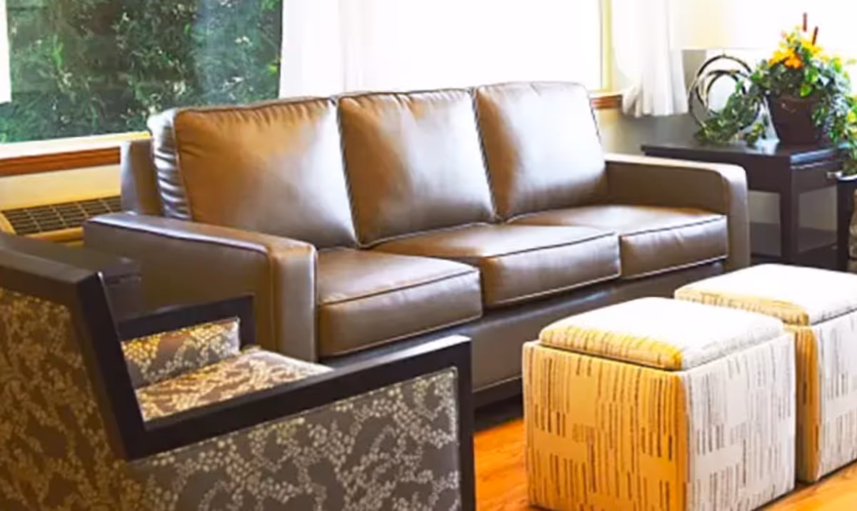 Bright seating area with a brown leather sofa, patterned armchair, two upholstered ottomans, and side tables with plants.