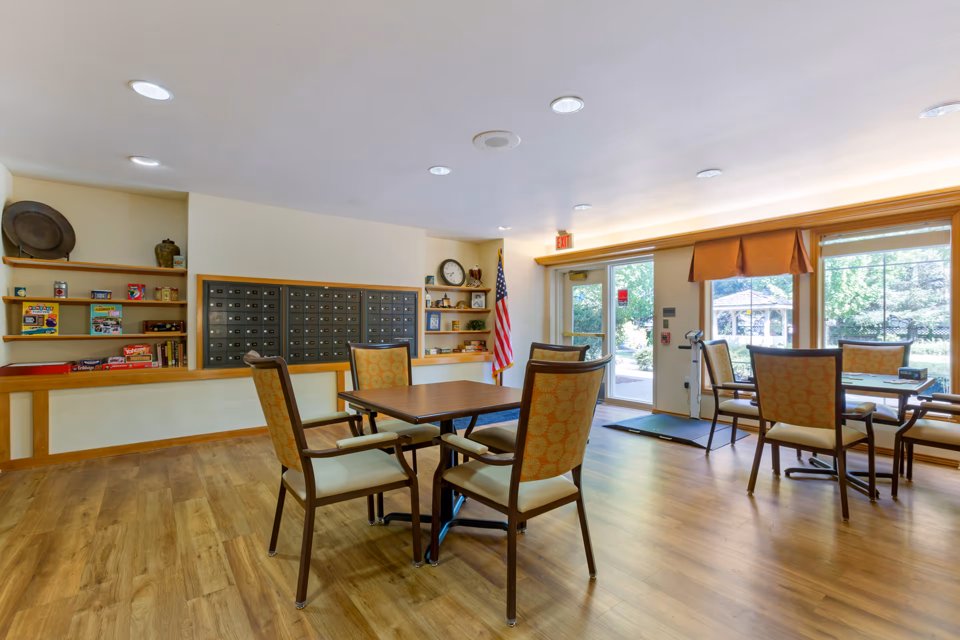 A bright room with wooden flooring featuring two tables each surrounded by four cushioned chairs. The room has a wall with multiple mailboxes and shelves holding various decorative items and board games. There is an American flag near a glass door that leads outside to a garden area with a gazebo.