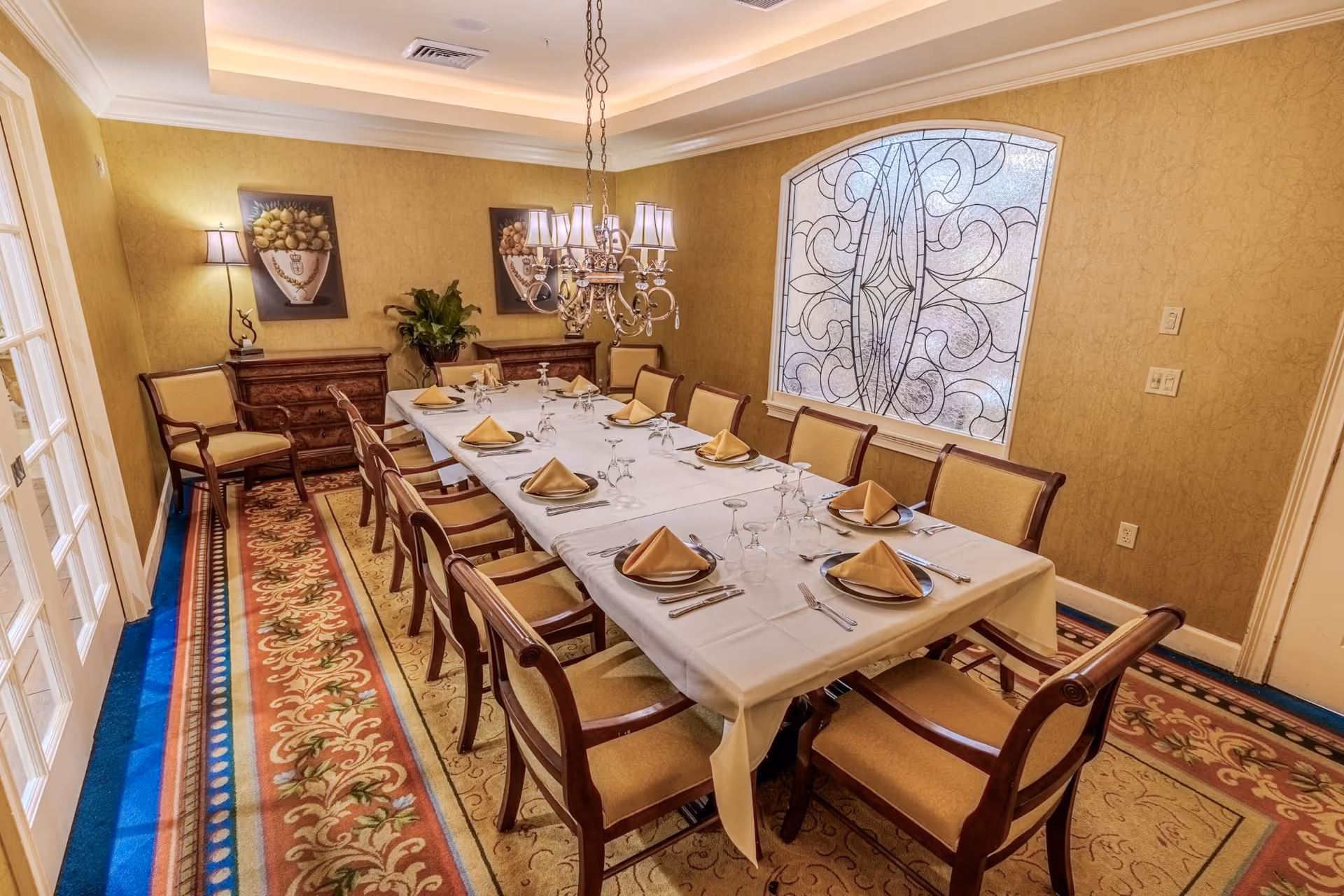 Elegant dining room with a long set table, chandelier, upholstered chairs, and a decorative stained-glass window.