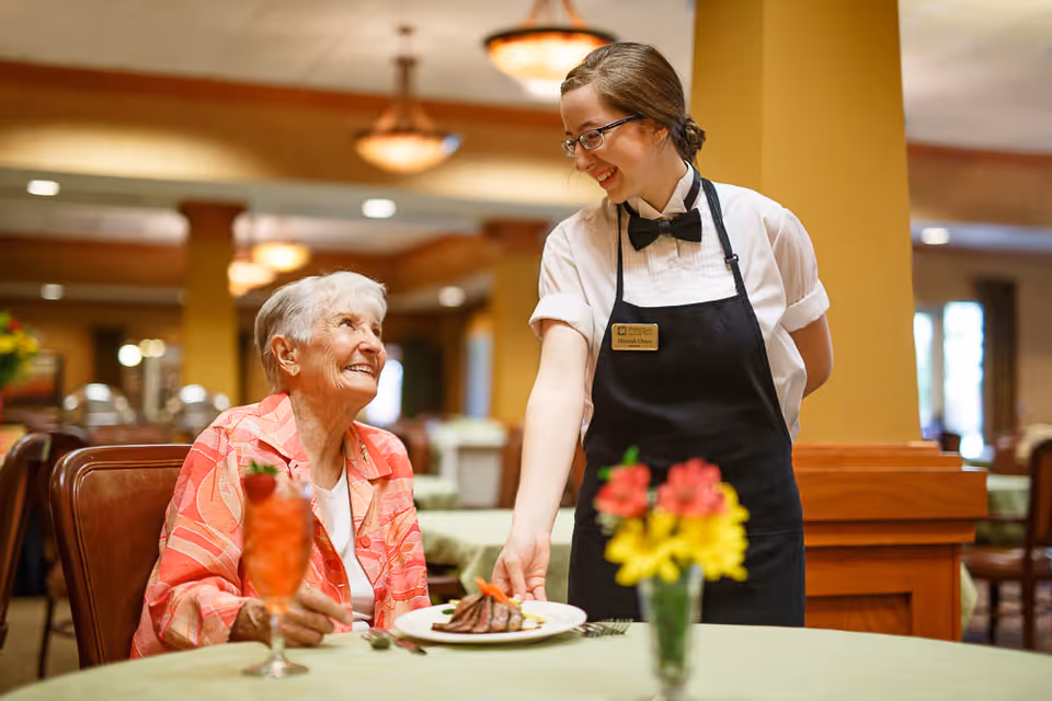 An elderly woman sitting at a dining table in a restaurant setting, smiling and looking up at a waitress who is serving a plate of food. The waitress is wearing glasses, a white shirt, black apron, and a bow tie. The table has a flower vase with yellow and red flowers and a red drink in a glass.