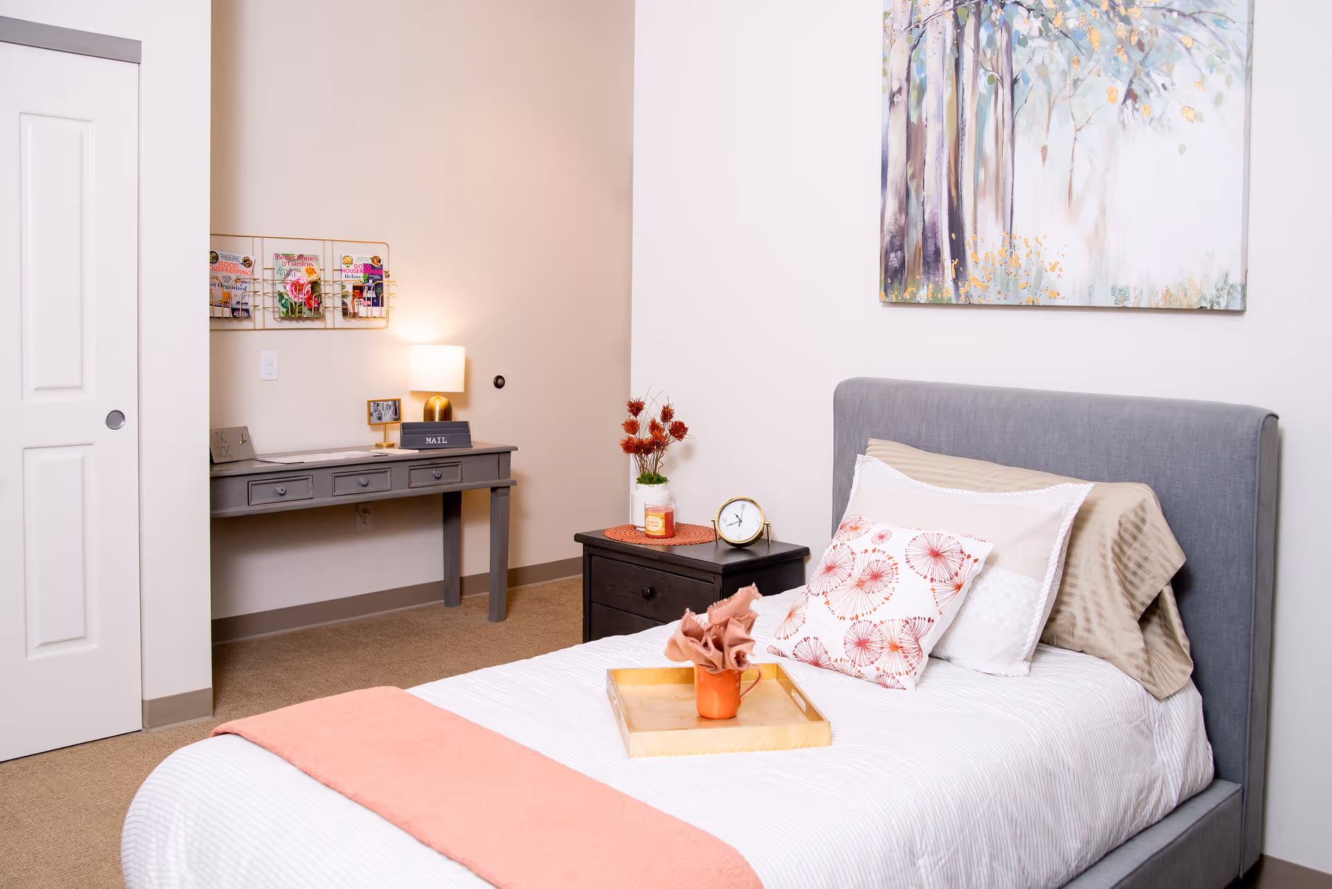 A neatly made single bed with a gray upholstered headboard, white bedding, and decorative pillows in a softly lit bedroom. A wooden tray with a small orange vase and napkin sits on the bed. Next to the bed is a dark nightstand with a clock, a candle, and a small plant. On the wall above the bed is a colorful painting of trees. In the background, there is a gray desk with a lamp, mail organizer, and wall-mounted magazine racks. The room has beige carpet and light-colored walls.