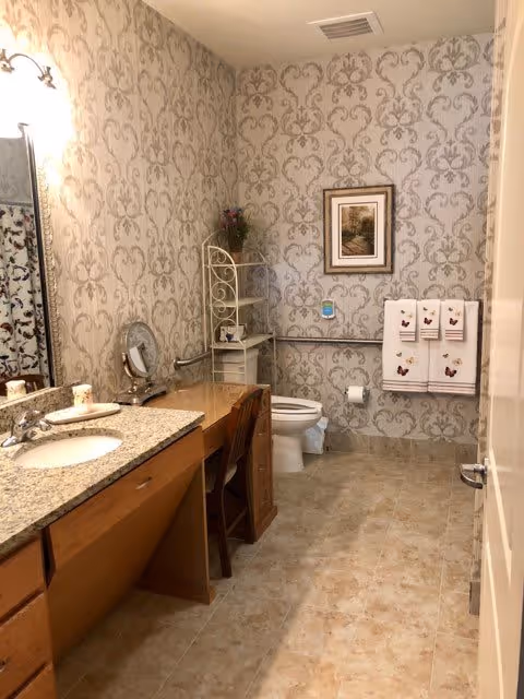A bathroom with beige patterned wallpaper, a granite countertop with a sink, a wooden vanity with a chair, a toilet with grab bars, a white metal shelving unit with decorative items, a framed picture on the wall, and three white towels with butterfly designs hanging on a towel rack.