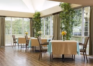 Bright dining room with tables covered in tablecloths, chairs, vases of flowers, and large windows overlooking trees.