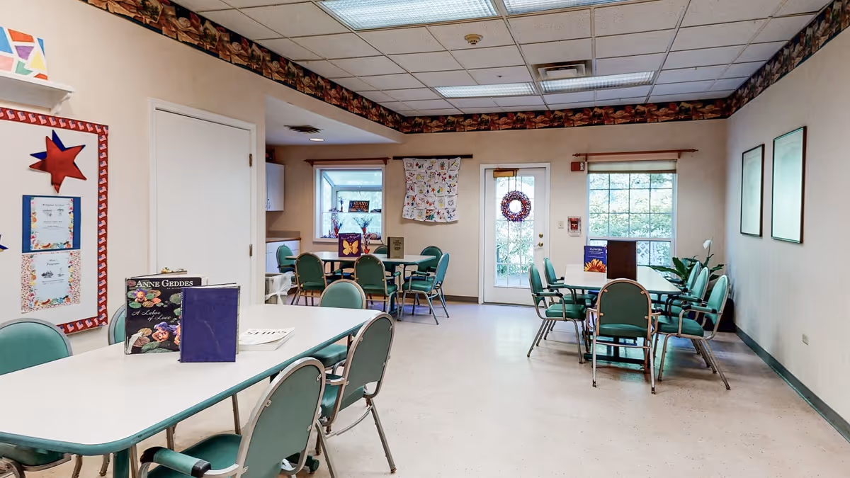 A well-lit common room with several tables and green chairs arranged around them. The room has beige walls with a decorative border near the ceiling, a bulletin board with colorful decorations on the left wall, and two windows and a glass door letting in natural light. There are books and pamphlets on the tables, and some framed pictures and plants are visible in the room.
