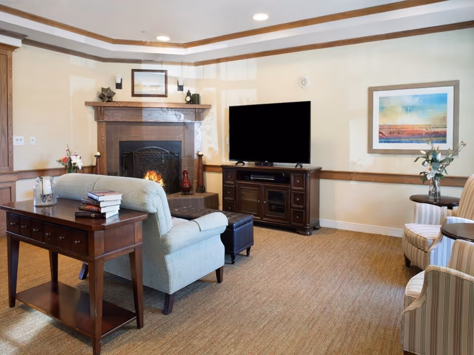 A cozy living room with a light blue upholstered sofa facing a dark wooden TV stand with a large flat-screen TV. There is a fireplace with a fire burning, framed artwork above it, and decorative items on the mantel. A wooden console table behind the sofa holds a stack of books and a candle. Two striped armchairs with a small round table and a vase with flowers are on the right side. The room has beige walls with wooden trim and a carpeted floor.