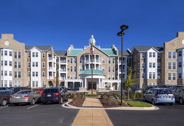 Front exterior view of a large senior living facility building with multiple floors, a central entrance with a green roof and columns, an American flag in front, and a parking lot with several cars under a clear blue sky.