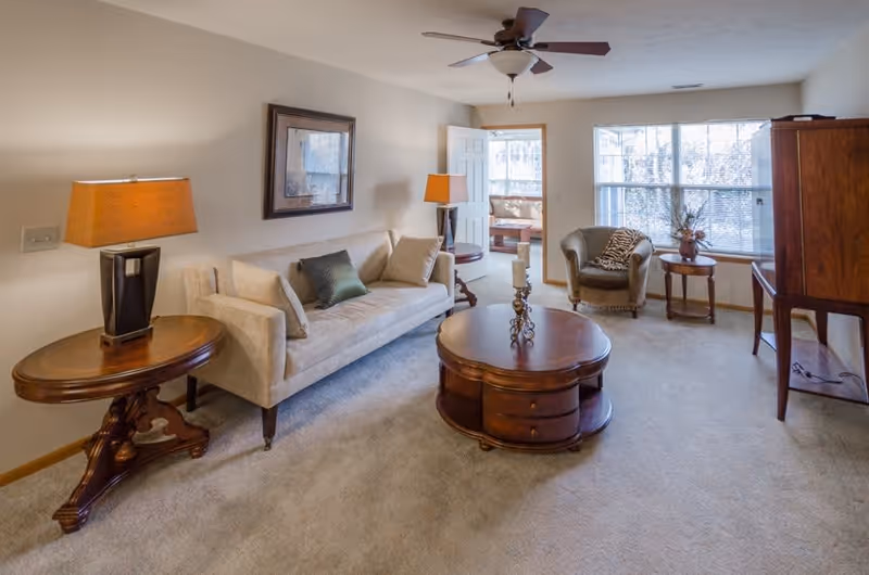 A cozy living room with a beige sofa adorned with cushions, a round wooden coffee table with decorative candles, a side table with a lamp, an armchair with a throw blanket, a large window letting in natural light, and a wooden cabinet. The room has carpeted flooring and a ceiling fan with lights.