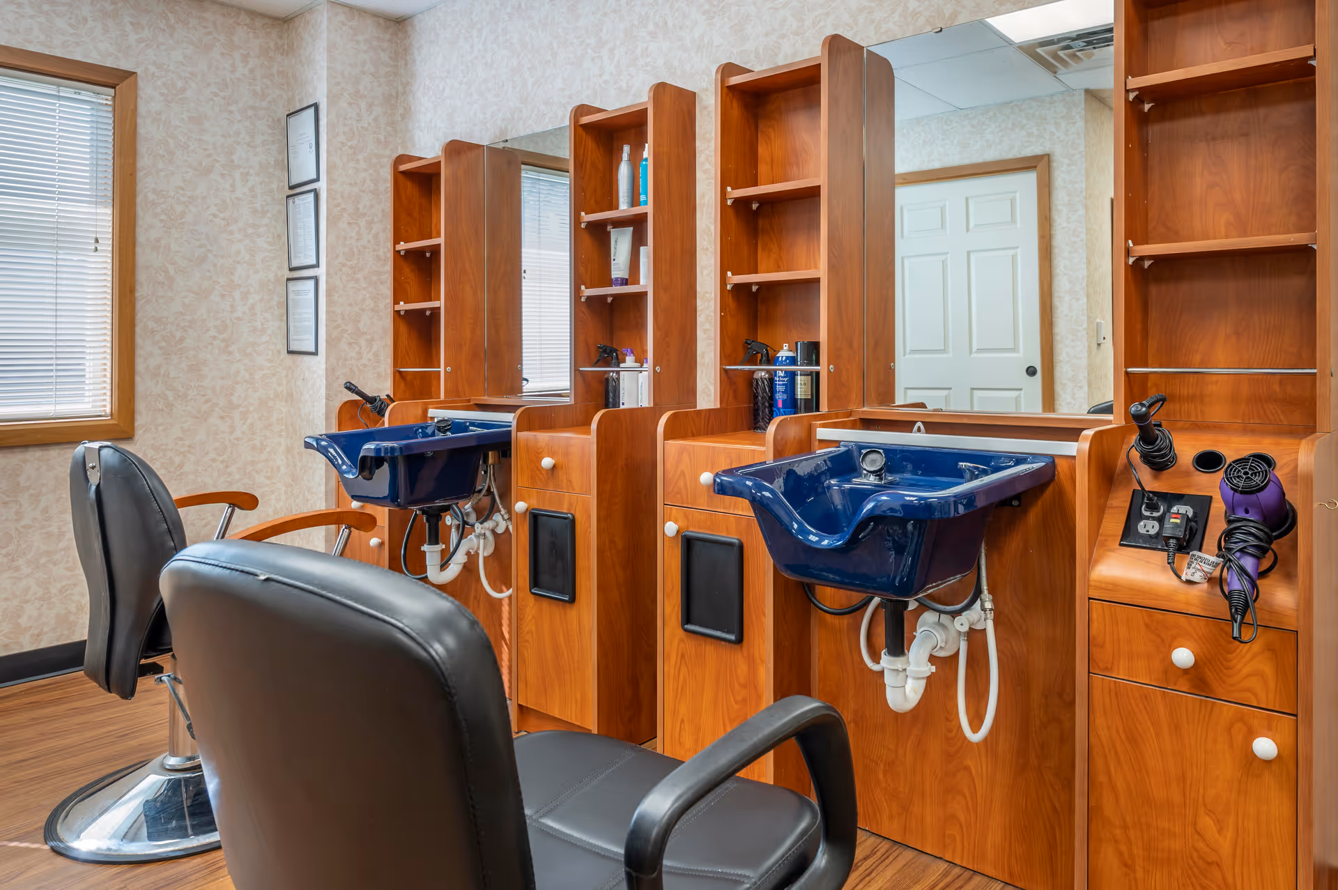 Interior view of a hair salon area with two black salon chairs facing wooden stations equipped with blue wash basins, mirrors, shelves, and hair styling tools including a hair dryer. The room has a window with blinds and a door in the background.