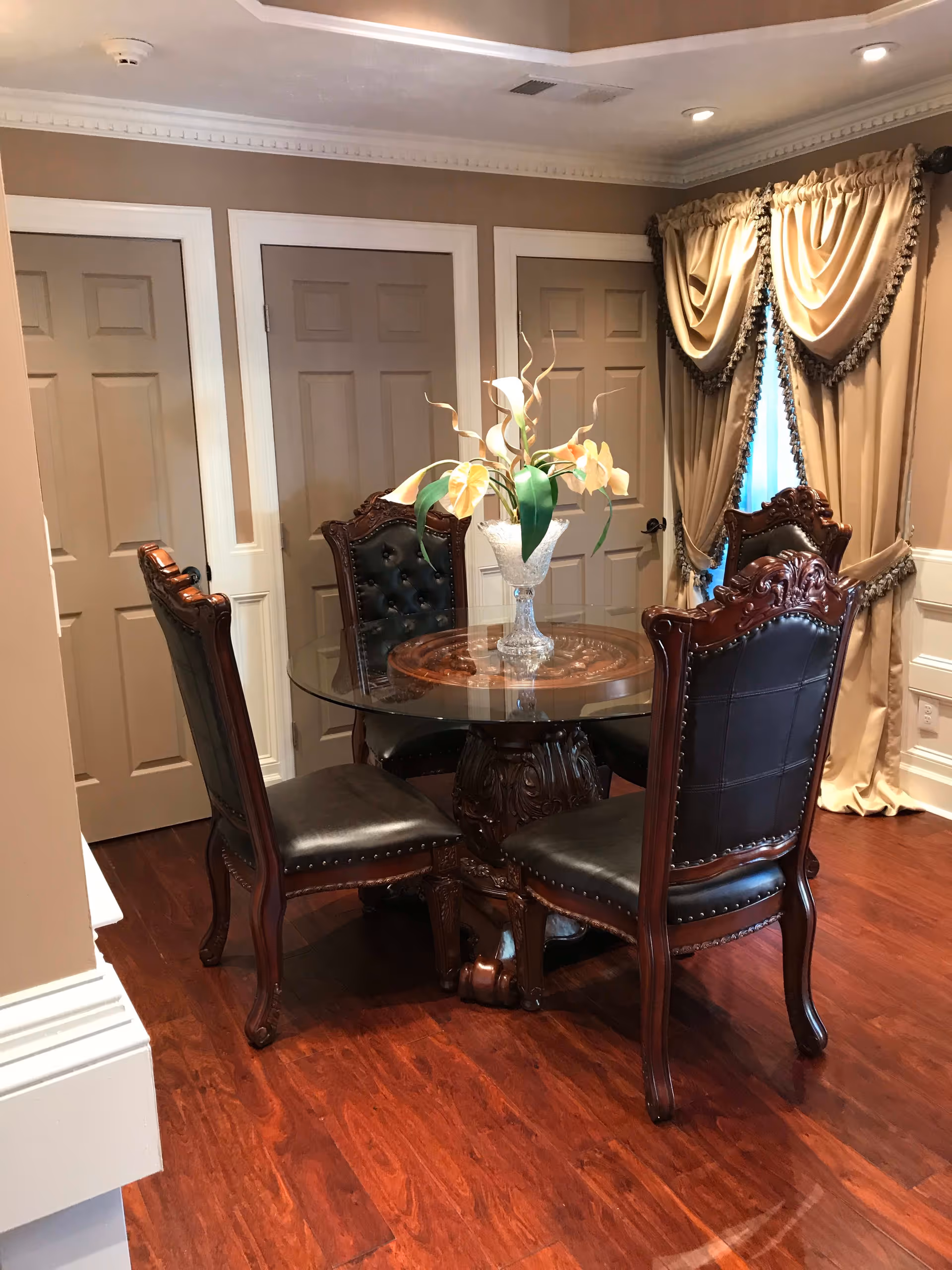 A small dining area with a round glass-top table and four ornate wooden chairs with black leather upholstery. A decorative vase with flowers is placed in the center of the table. The room has wooden flooring, beige walls, three closed doors, and a window with gold curtains and tassels.