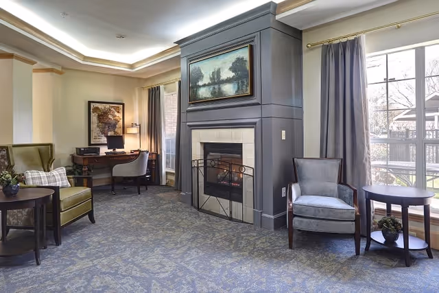 Bright seating area in a senior living lounge featuring a central fireplace, armchairs, side tables, a desk, and large windows.