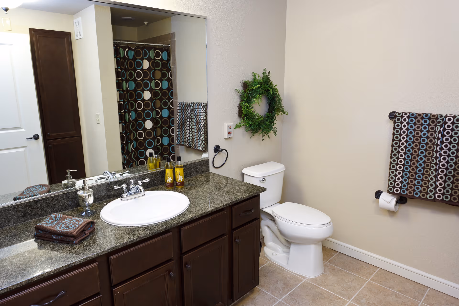 A clean bathroom featuring a white toilet, a dark granite countertop with a white sink, and dark wooden cabinets. There is a large mirror above the sink reflecting a shower curtain with a circular pattern in brown, blue, and white. A matching towel hangs on a towel rack next to the toilet, and a green wreath is mounted on the wall above the toilet. The floor is tiled in a light beige color.