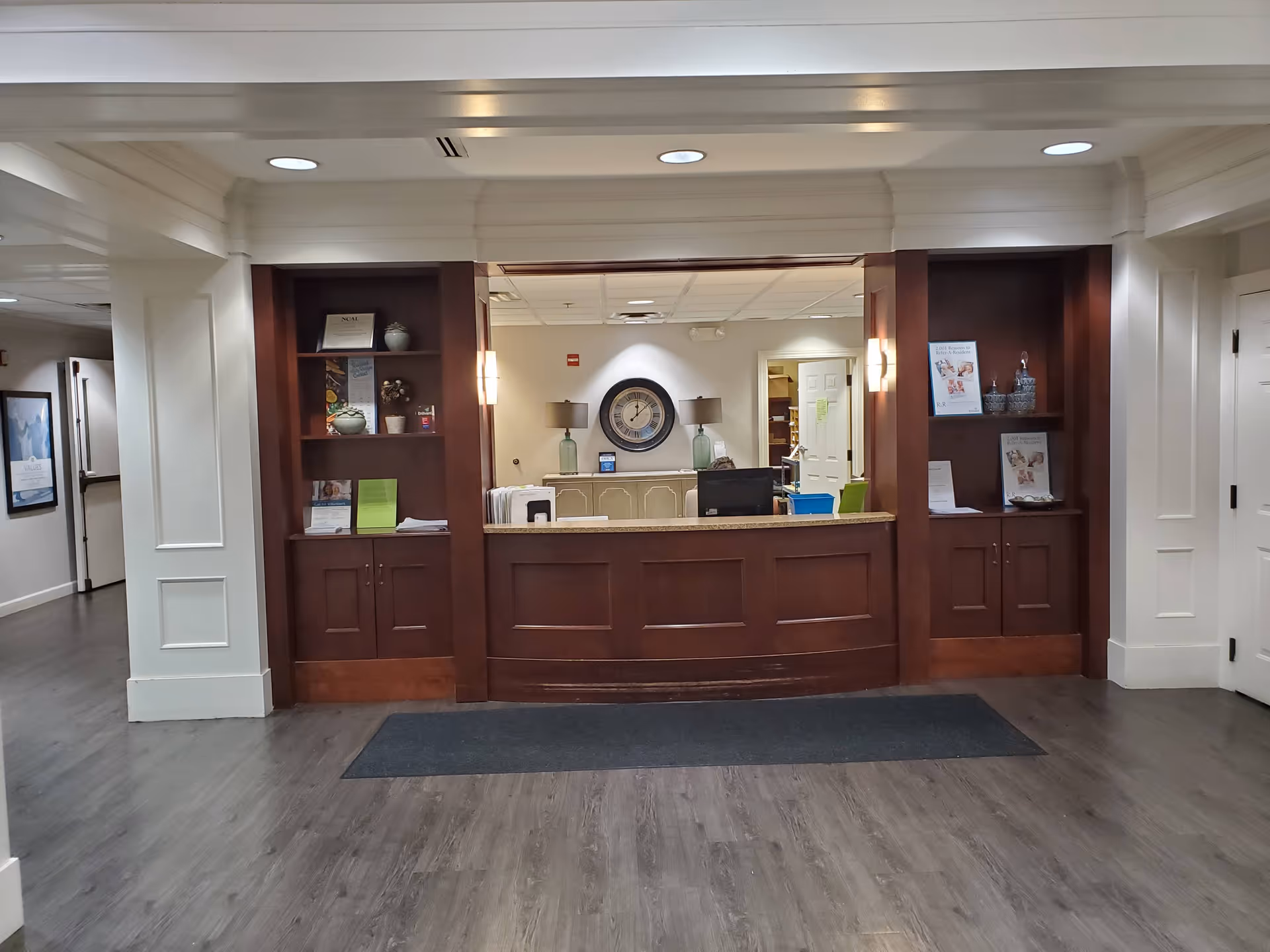 Reception area with a wooden front desk and built-in shelves on either side. The shelves hold decorative items, brochures, and informational materials. Behind the desk is a wall with a large clock and two lamps on a sideboard. The floor is wood with a dark rug in front of the desk, and the ceiling has recessed lighting.