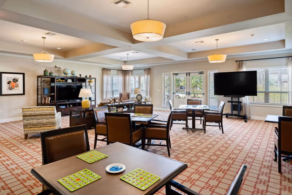 Well-lit senior living common room with tables and chairs set for games, a TV, shelving, and large windows.