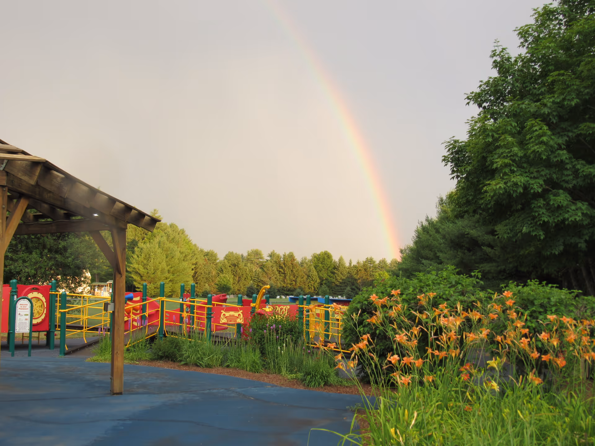Outdoor playground with colorful play equipment, a wooden shelter, flowering plants and trees beneath a rainbow.