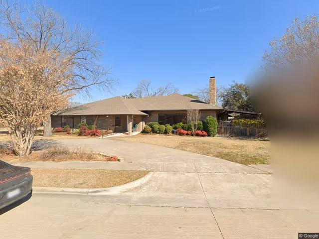 Single-story brick building with a sloped roof, surrounded by a lawn with bushes and trees, under a clear blue sky. A driveway leads up to the entrance, and a car is partially visible on the left side.