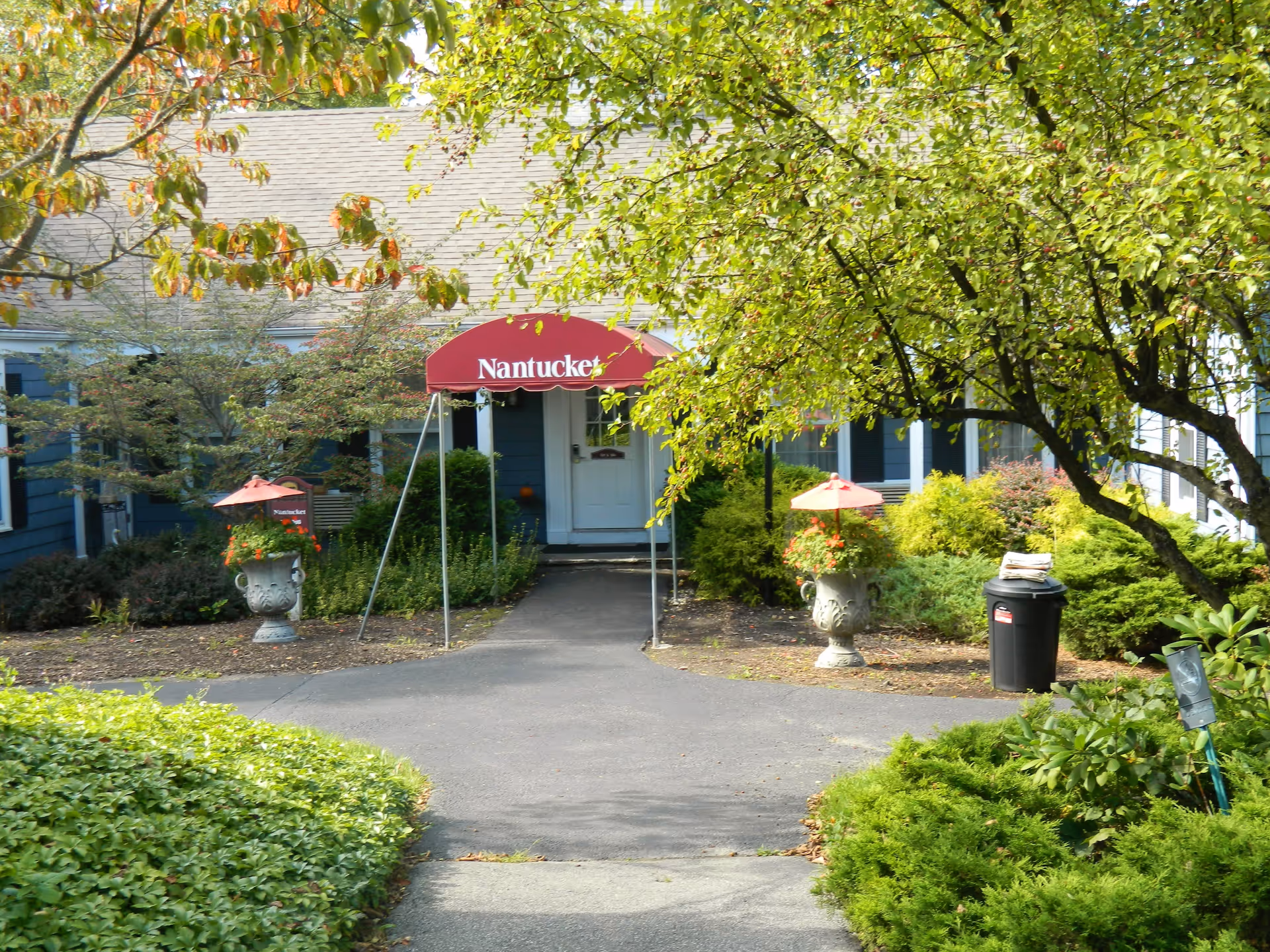 Entrance to a building with a red awning labeled 'Nantucket' above the door, surrounded by green bushes and trees with some autumn leaves. There are two large decorative planters with red umbrellas and flowers on either side of the walkway leading to the door.
