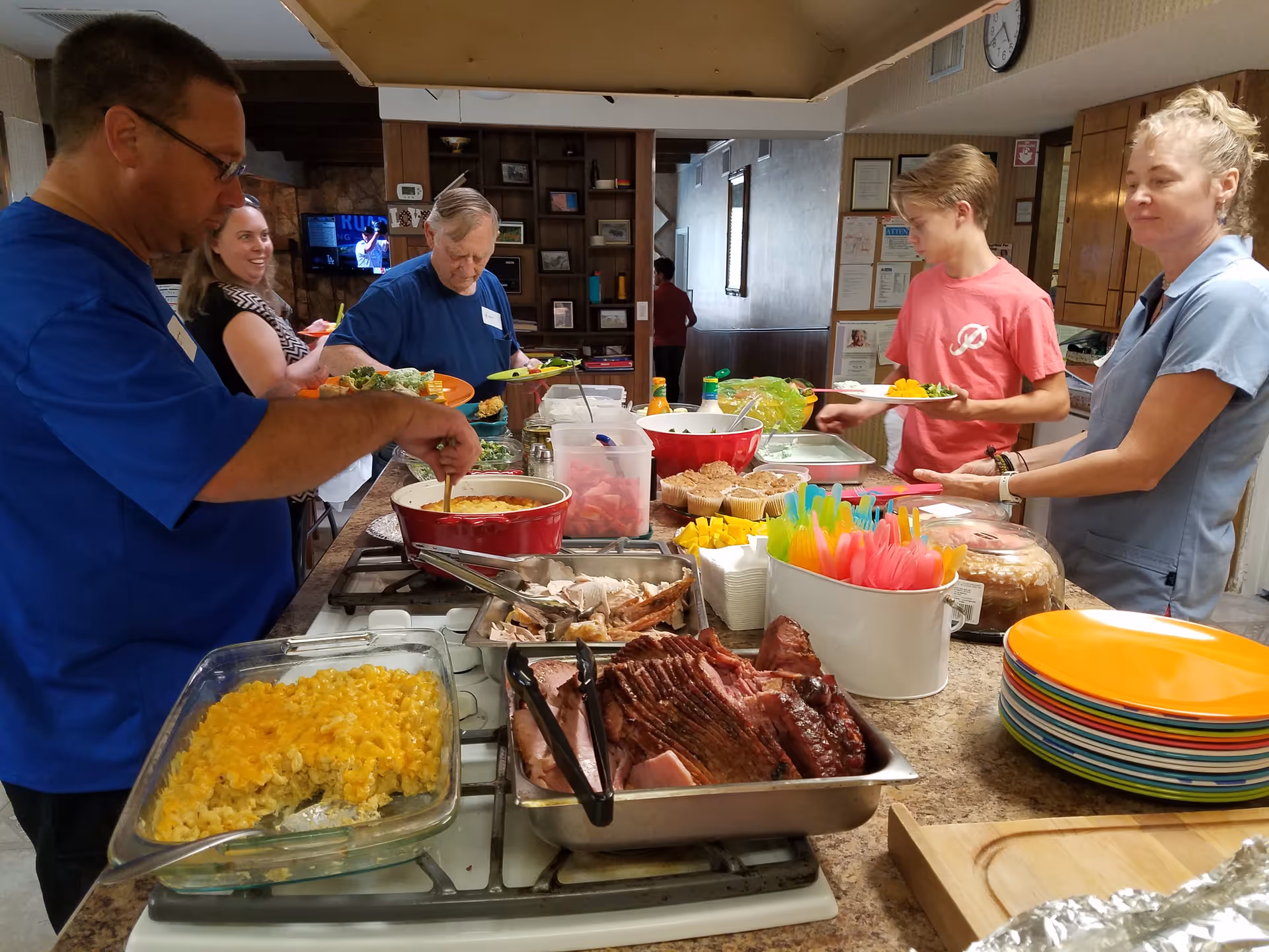 Several people serving themselves from a buffet of hot dishes and salads on a counter in a communal dining/kitchen area.