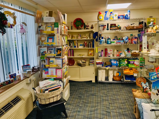 Interior view of a small shop or supply room with shelves stocked with various household and personal care products, greeting cards on a rotating display rack, and decorative items on shelves. The room has a window with vertical blinds, a carpeted floor, and fluorescent ceiling lights.