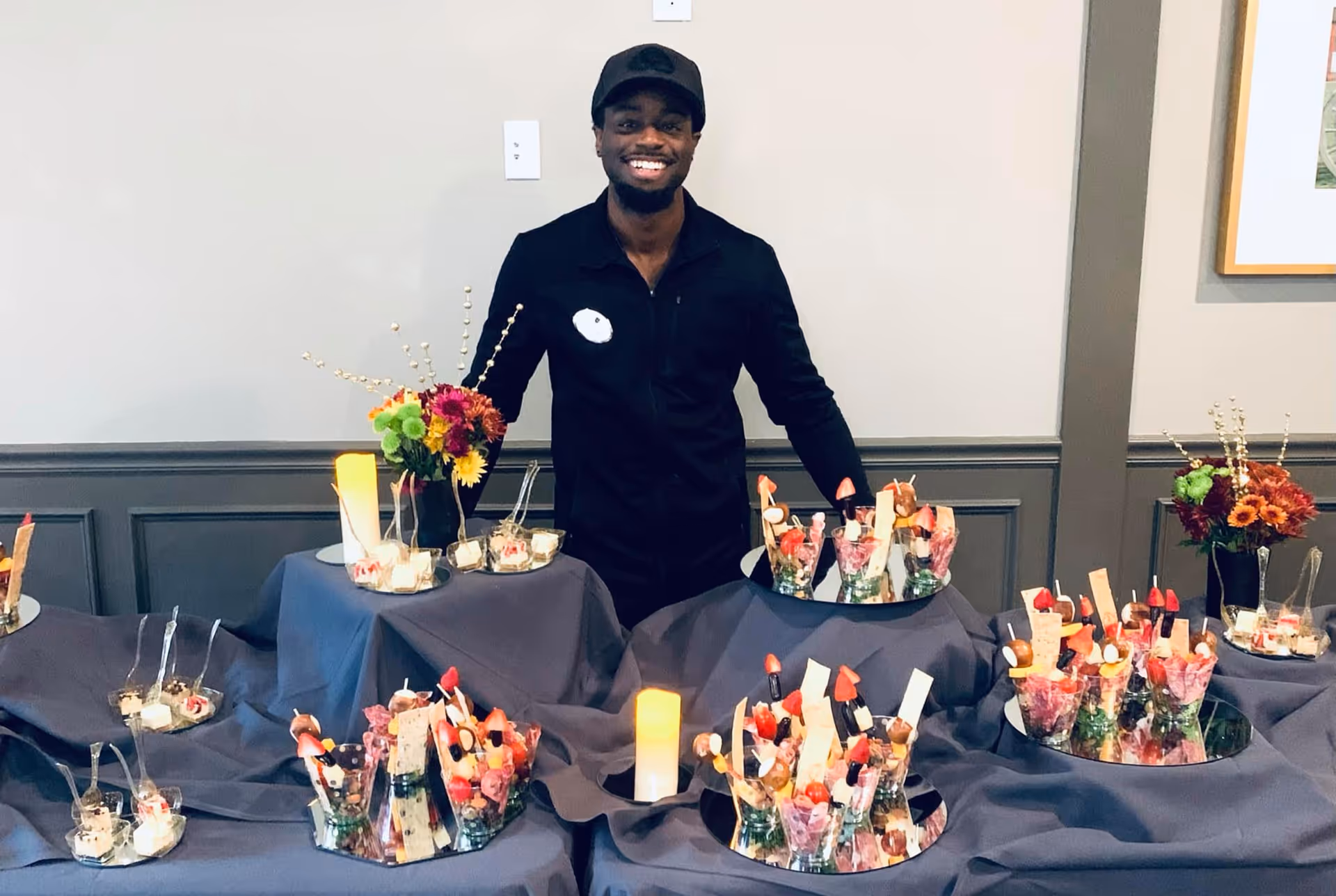 A smiling man stands behind a table arranged with individual dessert cups and floral centerpieces in a dining/event space.