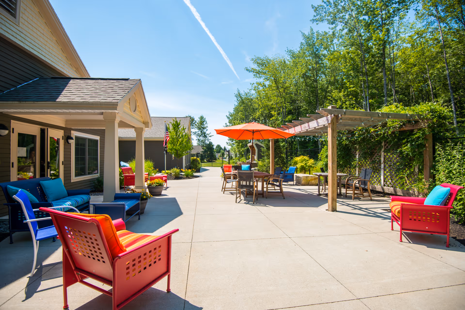 Outdoor patio area at The Highlands with colorful chairs and cushions, a table with an orange umbrella, a wooden pergola with climbing plants, and a clear blue sky with trees in the background.